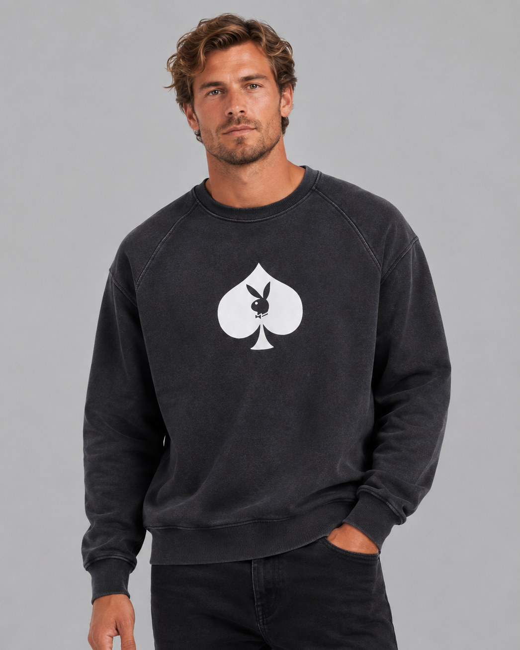 A man wearing a faded black crewneck sweatshirt with a large white Playboy bunny and spade logo on the chest, paired with black jeans, against a solid gray studio background.