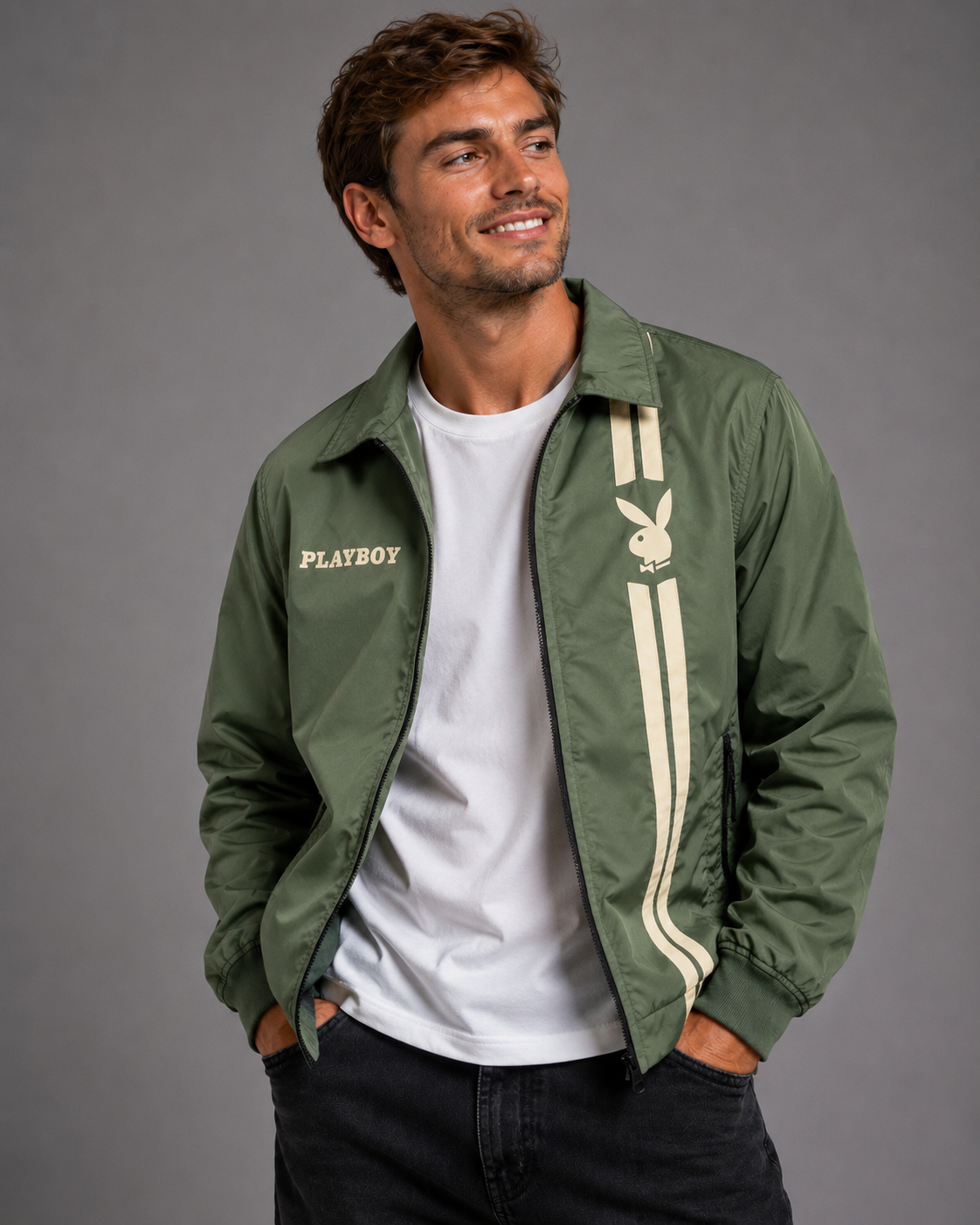 A man wearing an olive green Playboy bomber jacket over a white t-shirt and dark jeans, standing against a solid gray studio background.