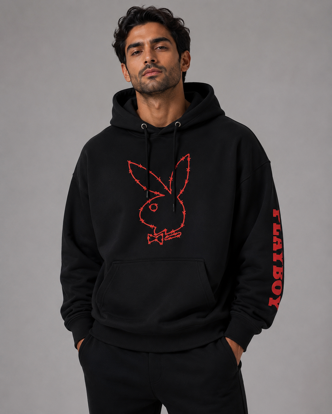 A man wearing a black pullover hoodie with a red Playboy bunny logo on the chest and red 'PLAYBOY' text down the sleeve, posed against a neutral gray studio background.