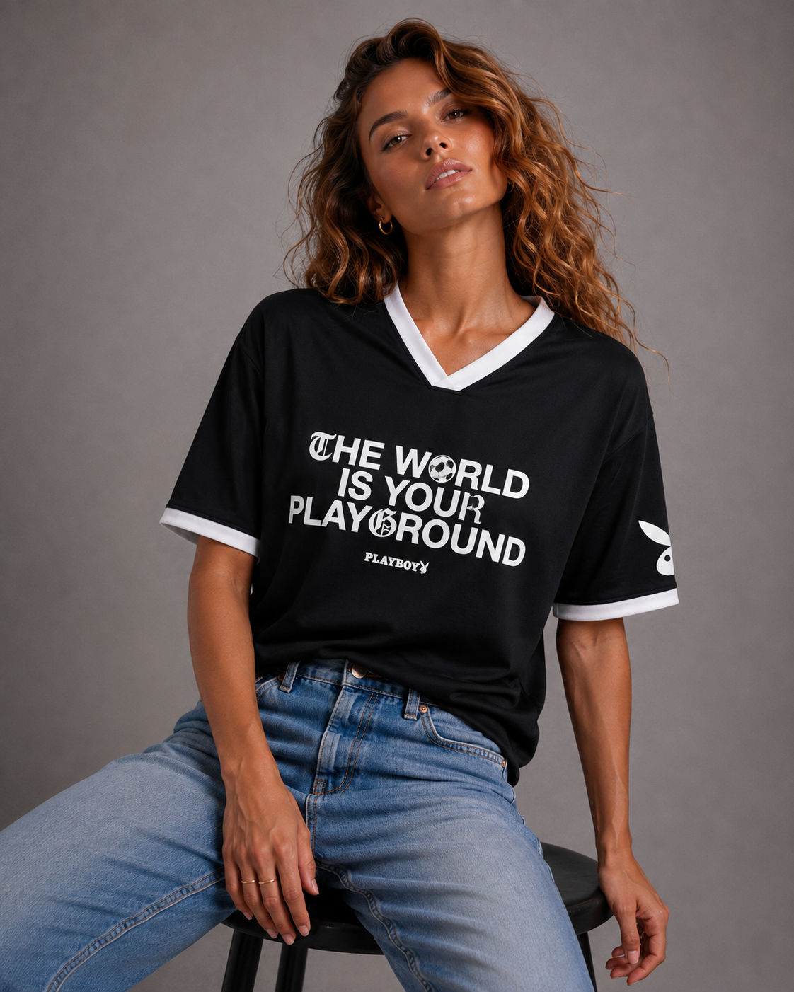 A woman wearing a black Playboy short-sleeve V-neck t-shirt with white trim and a white graphic print that reads 'THE WORLD IS YOUR PLAYGROUND' and 'PLAYBOY'. She is sitting on a stool against a neutral gray studio background. #Women