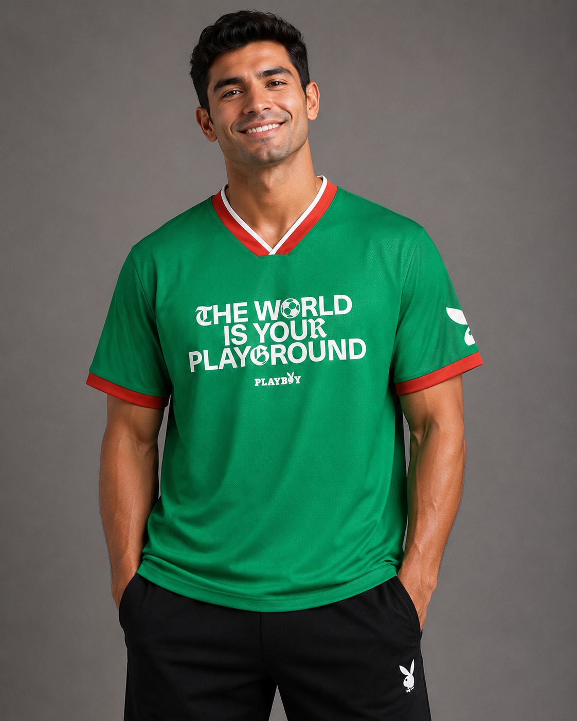 A man wearing a green Playboy short-sleeve soccer jersey with red and white trim, featuring the slogan 'THE WORLD IS YOUR PLAYGROUND' on the front, posed against a solid gray studio background.