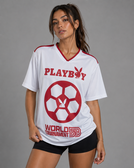 A woman modeling a white short-sleeve V-neck t-shirt with red Playboy branding and a red soccer ball graphic on the front, paired with black shorts against a solid gray studio background.