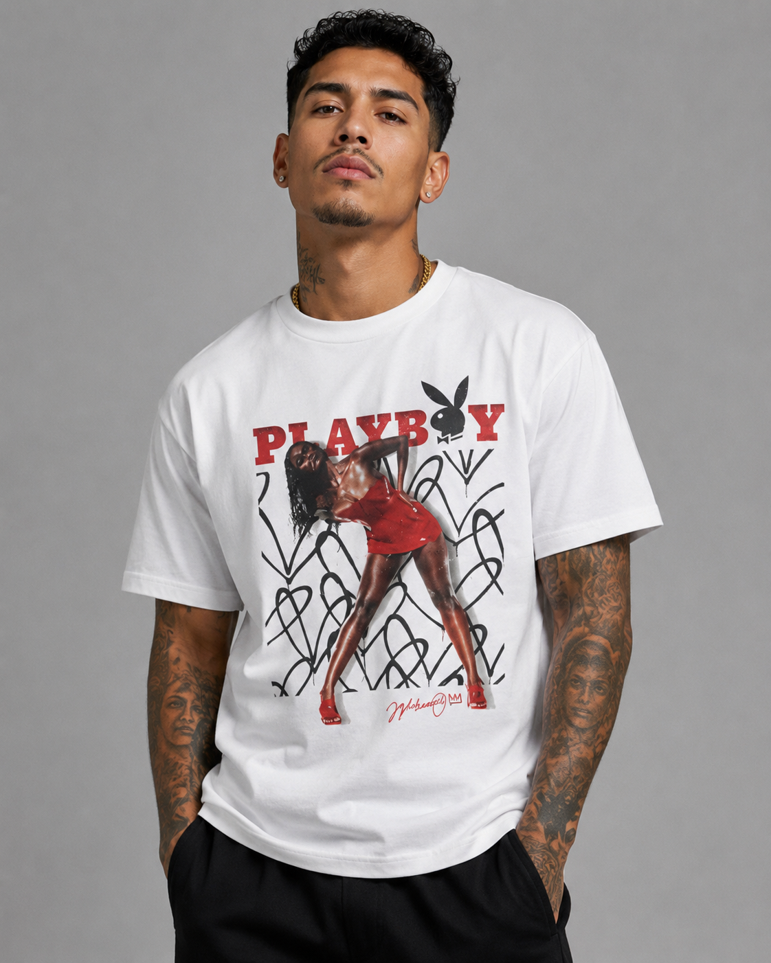 A man wearing a white short-sleeve graphic t-shirt featuring the red and black PLAYBOY logo and a stylized illustration of a woman, paired with black pants, against a solid gray studio background.