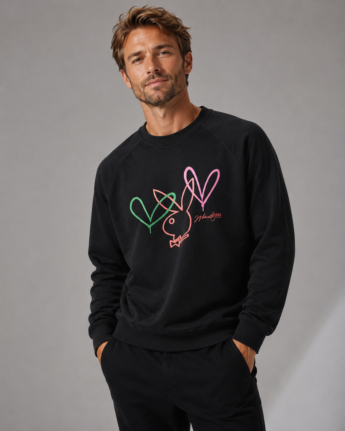 A man wearing a black crewneck sweatshirt with a colorful graphic of three hearts and a peace sign on the chest, paired with black sweatpants, against a neutral gray studio background.