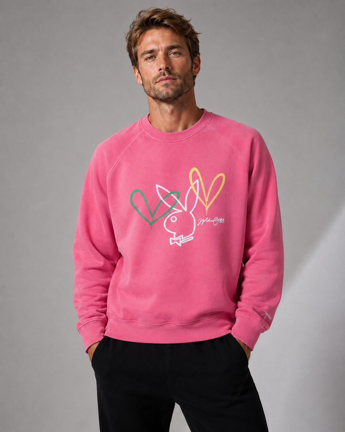 A man wearing a pink crewneck sweatshirt with a white, green, and yellow Playboy bunny and heart graphic on the front, paired with black pants, set against a plain gray studio background.