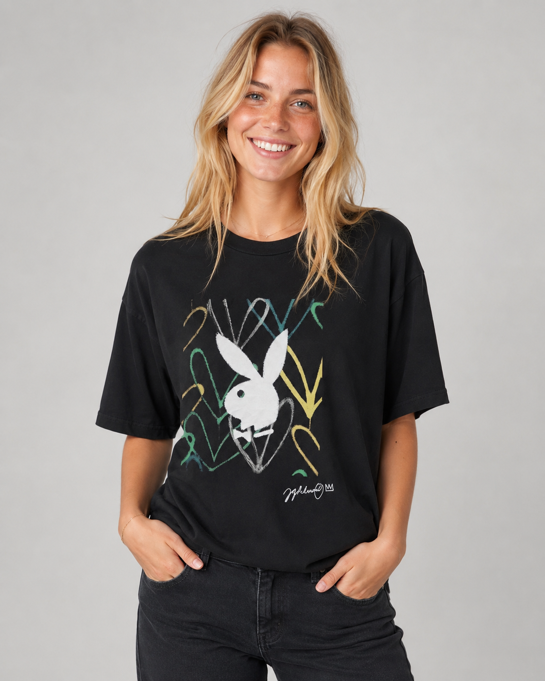 A woman wearing a black Playboy short-sleeve t-shirt with a white bunny graphic and colorful scribbled line details, paired with dark jeans, against a solid gray studio background. #Women