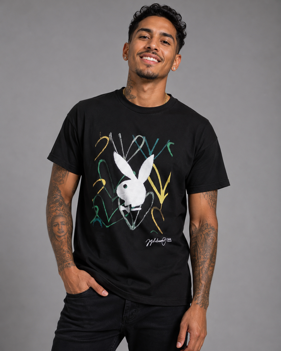 A man wearing a black graphic t-shirt featuring the white Playboy bunny logo surrounded by colorful graffiti-style lines, posed against a solid gray studio background.