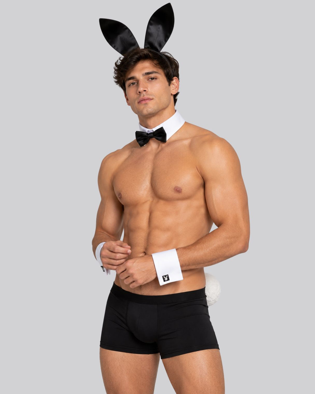 A shirtless man modeling a black bunny costume set with black bunny ears, a black bow tie, white cuffs, and black briefs against a solid gray background.