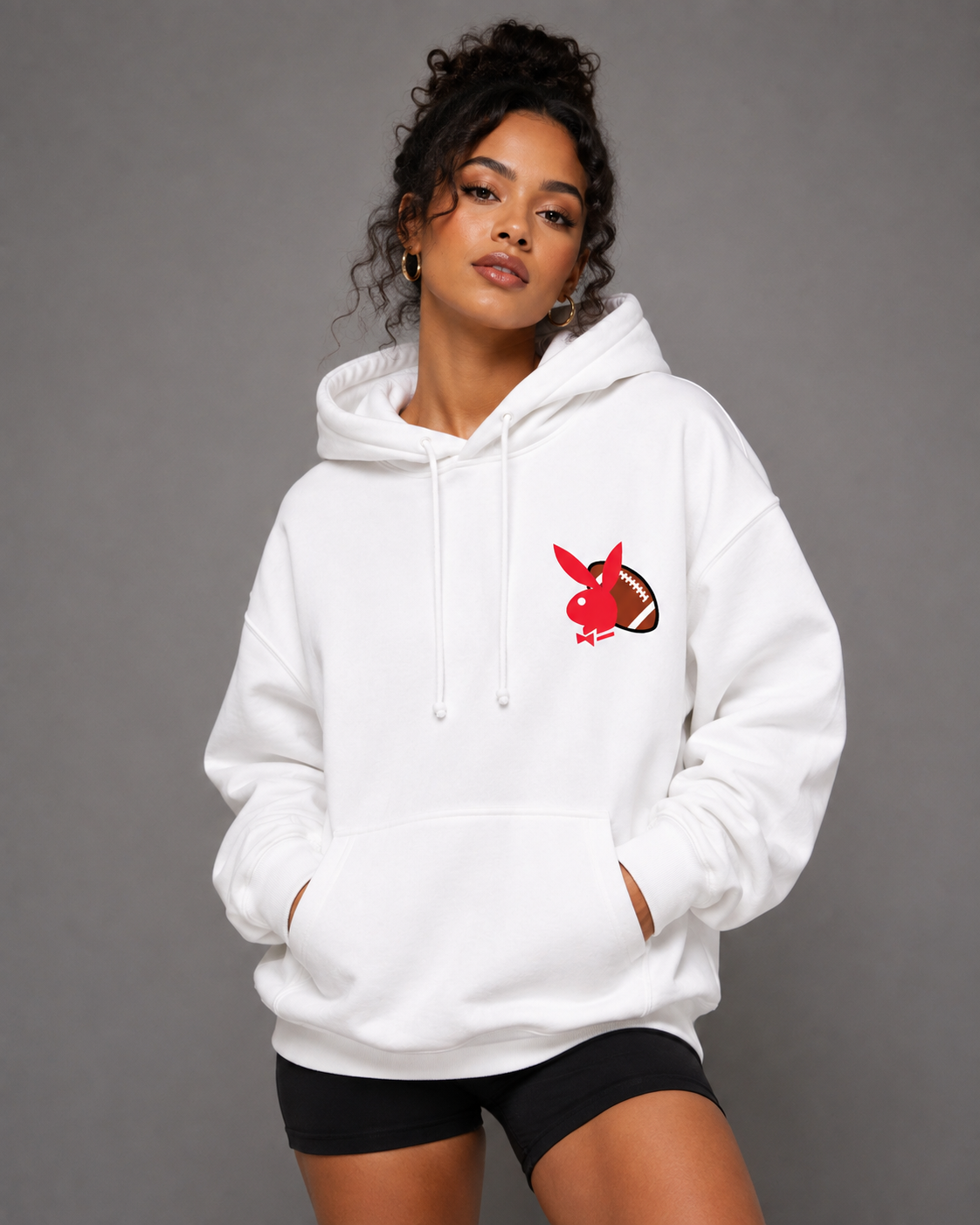 A woman modeling a white pullover hoodie with a red and brown rabbit graphic on the left chest, paired with black shorts, against a solid gray studio background.