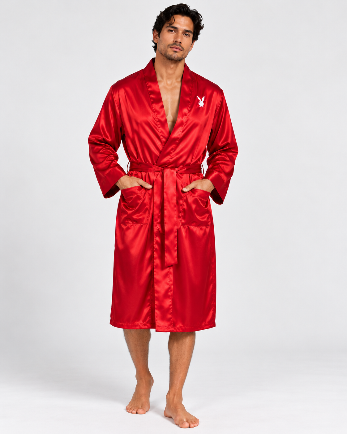 A man modeling a bright red satin bathrobe with a white Playboy bunny logo on the chest, standing against a light gray studio background.