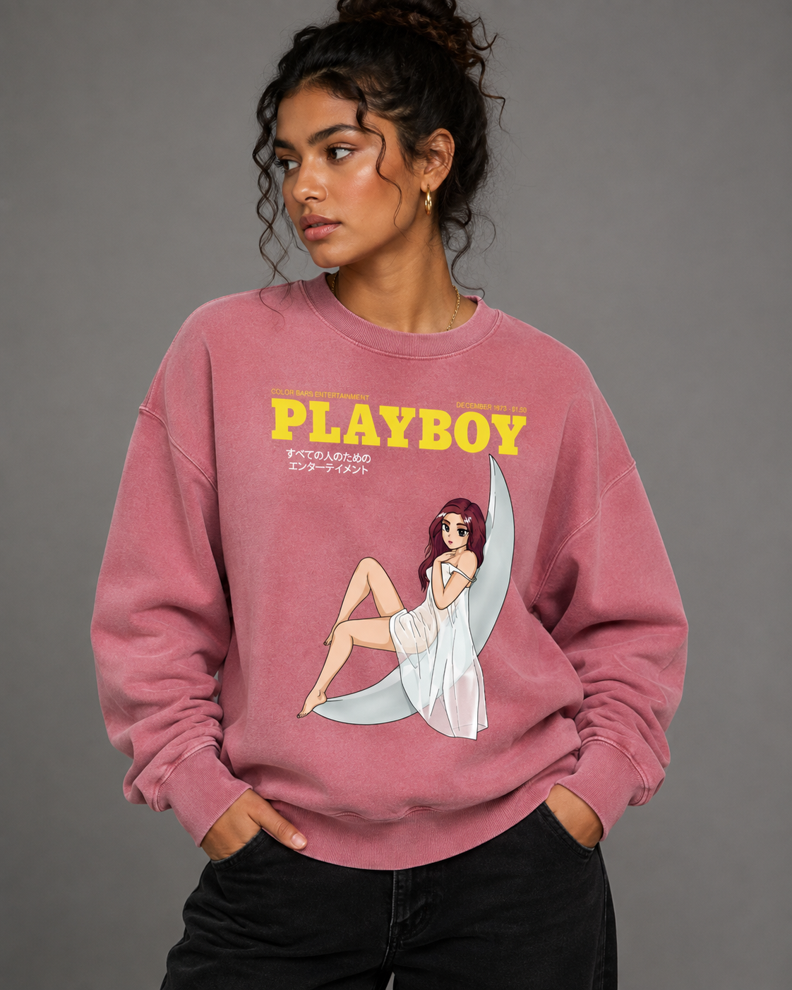 A woman wearing a pink crewneck sweatshirt with a yellow 'PLAYBOY' logo and a pin-up girl graphic on a crescent moon, paired with black jeans, against a solid gray studio background. #Streetwearwomen