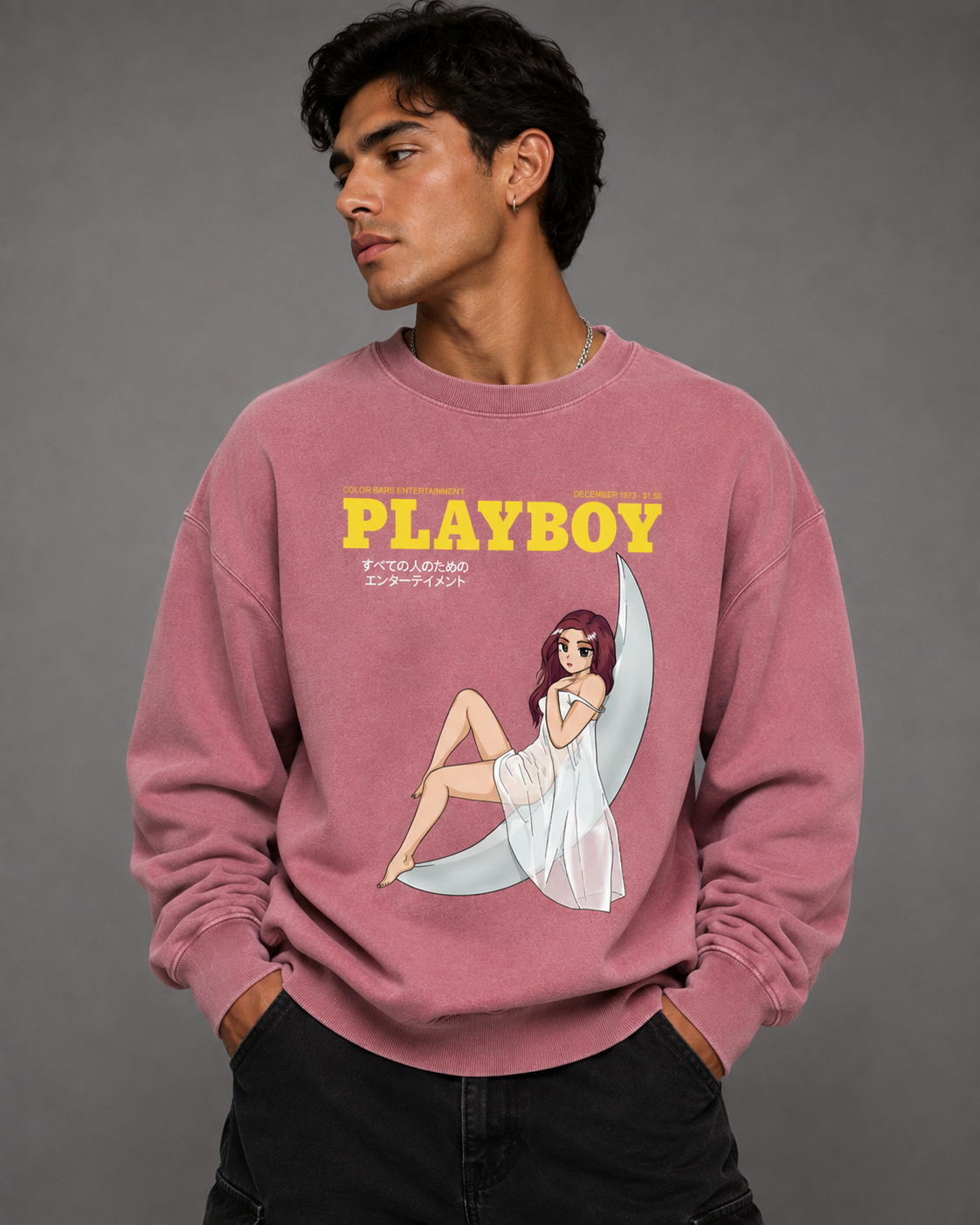 A man wearing a dusty rose pink crewneck sweatshirt with a yellow 'PLAYBOY' logo and a vintage-style pin-up illustration on the front, paired with black pants, against a solid gray studio background.