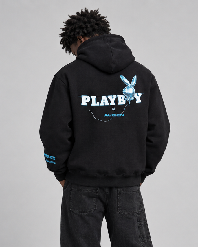 A model viewed from the back wearing a black PLAYBOY hoodie with a large white and blue graphic on the back, paired with black jeans and white sneakers, against a light gray studio background.