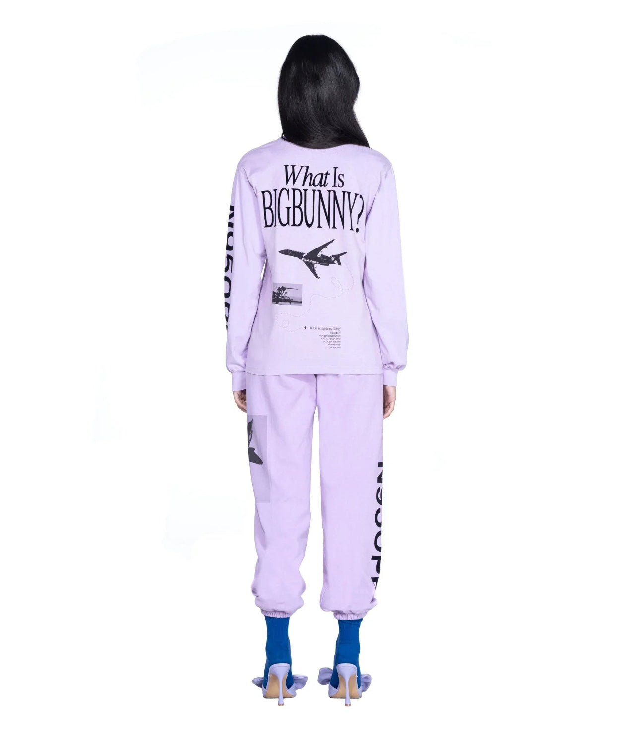 Woman wearing lavender long sleeve graphic top and matching joggers with airplane and text print, blue heels.