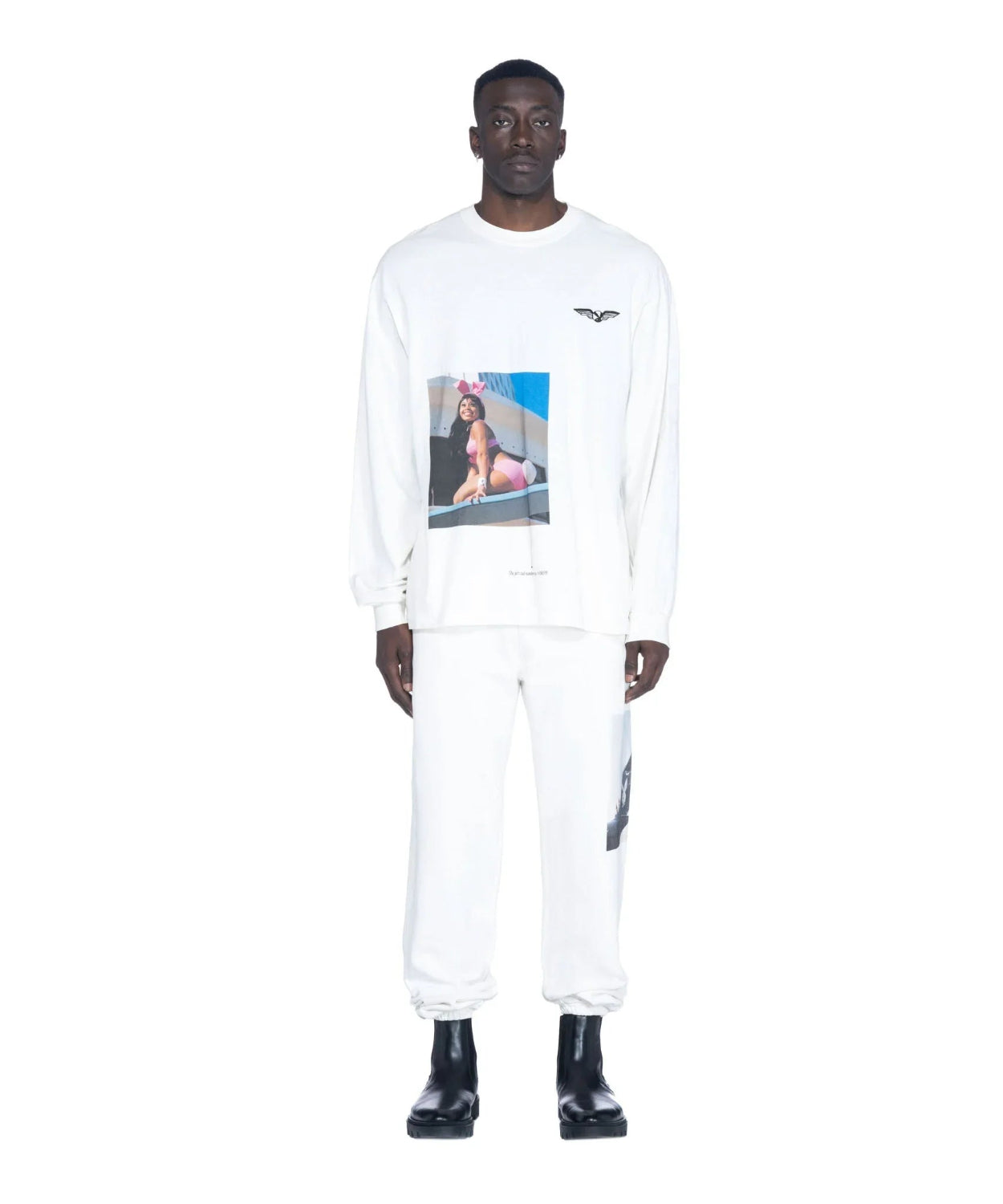 Man wearing white long sleeve T-shirt with aviation wings logo and graphic print, paired with white pants and black boots, standing against a plain background.