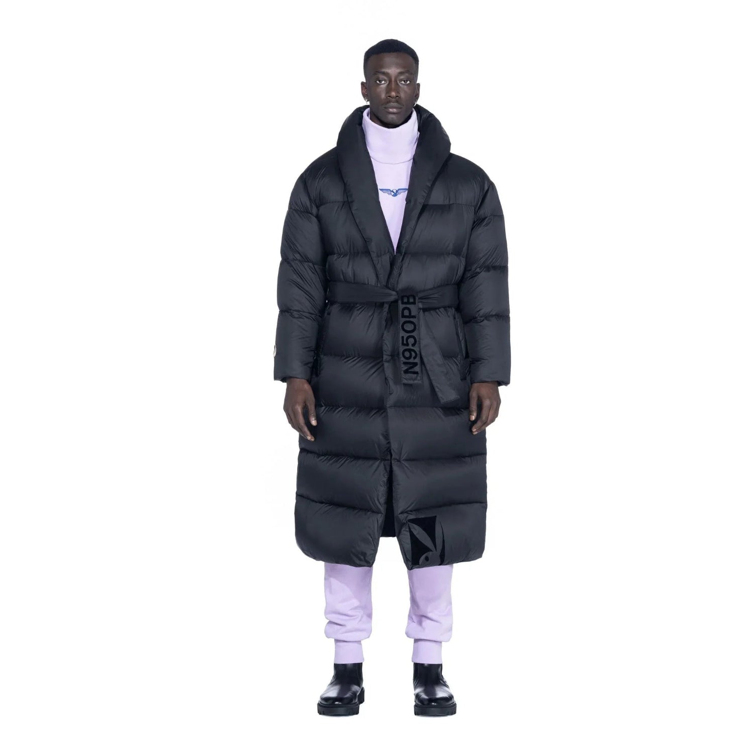 Man in black belted long puffer jacket, lilac pants, and black boots on white background