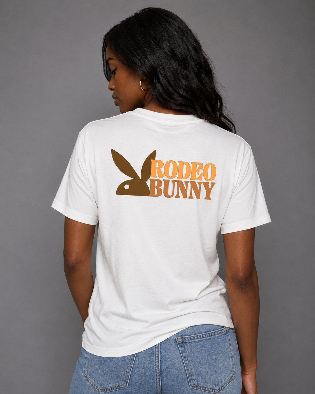 A woman shown from the back wearing a white short-sleeve t-shirt with a brown and orange graphic on the back. The graphic features the Playboy bunny head logo integrated with the text 'RODEO BUNNY'.