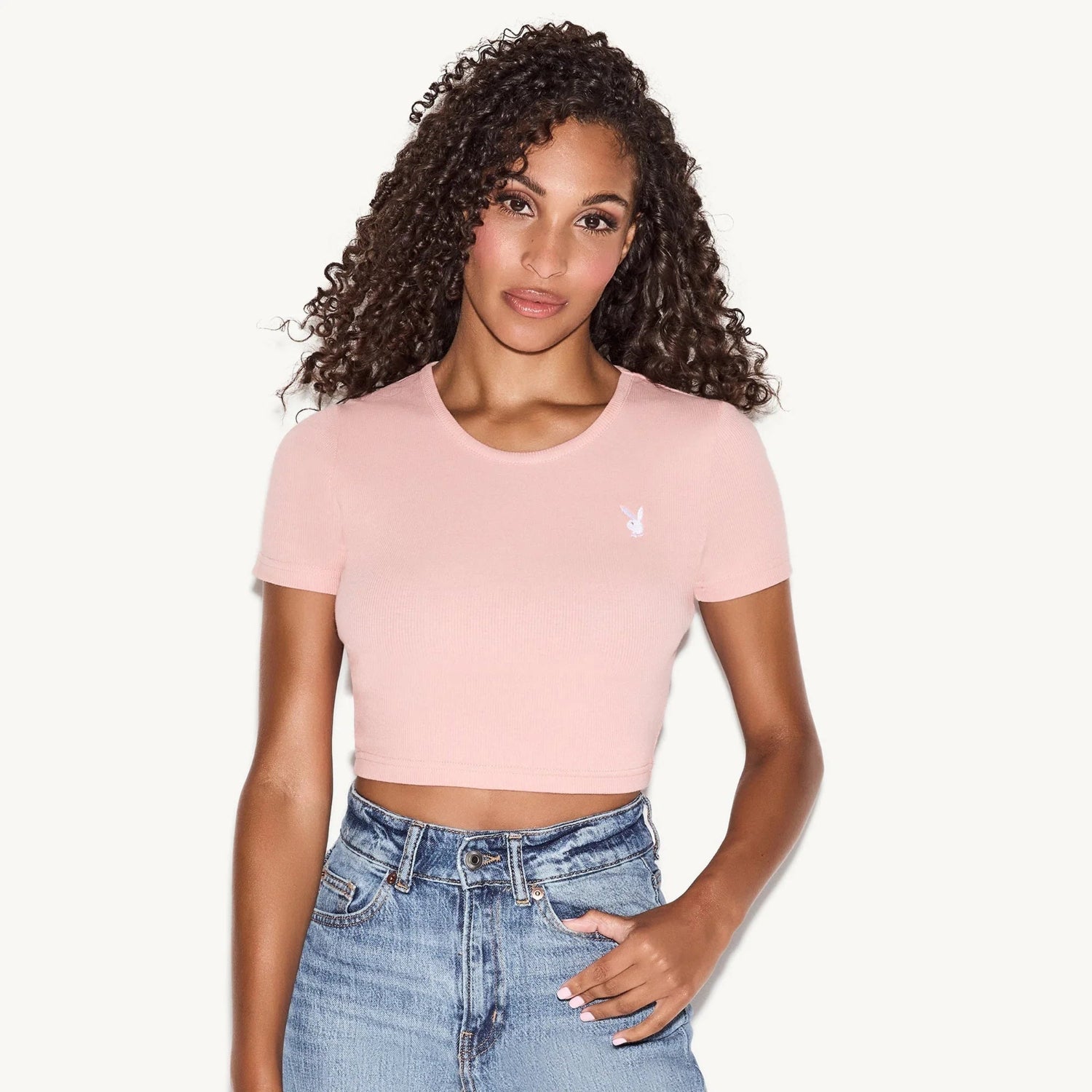 Woman wearing light pink cropped baby tee with small white logo and high-waisted jeans