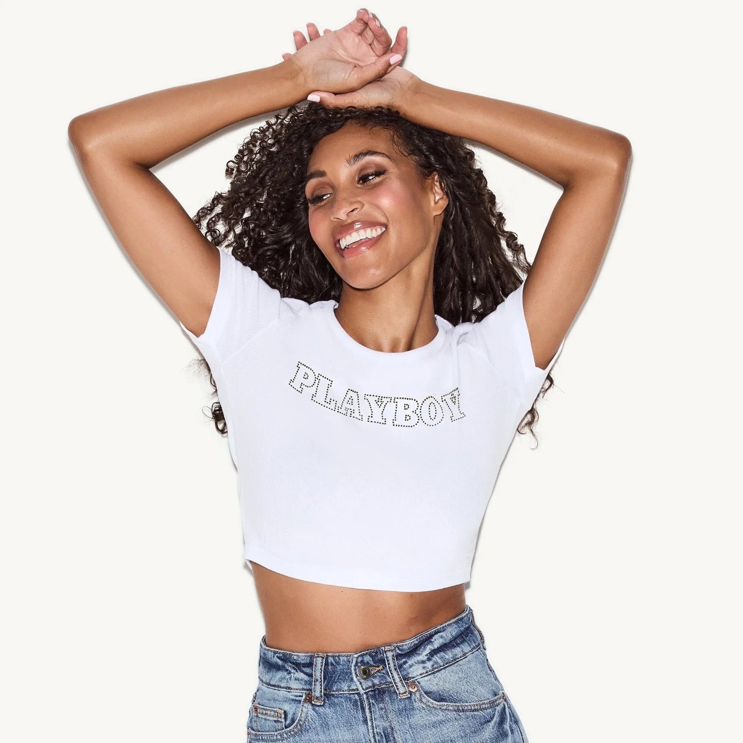 Woman wearing white rhinestone logo baby tee and blue denim jeans, posing with arms raised