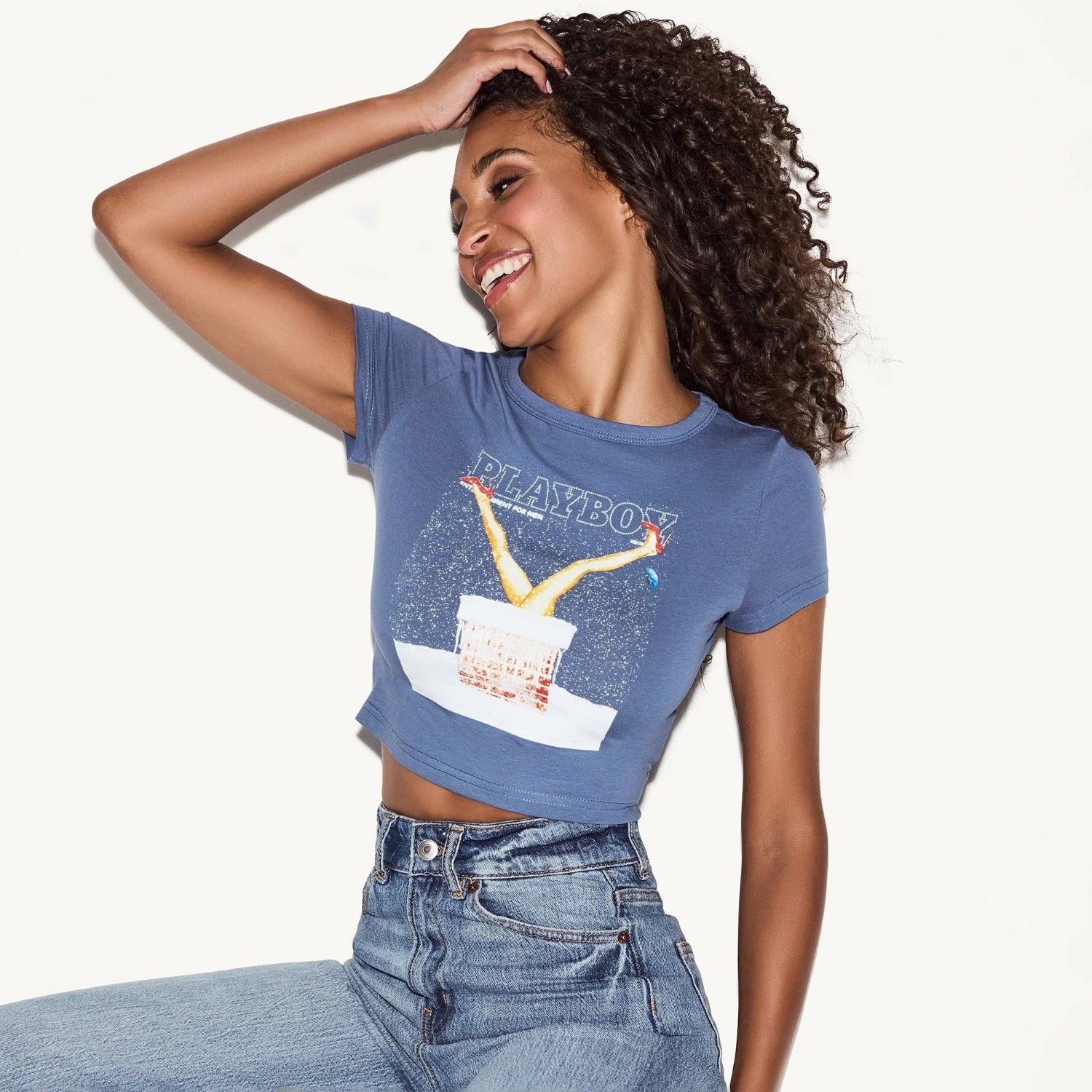 Smiling woman in blue graphic crop top and high-waisted jeans posing against white background