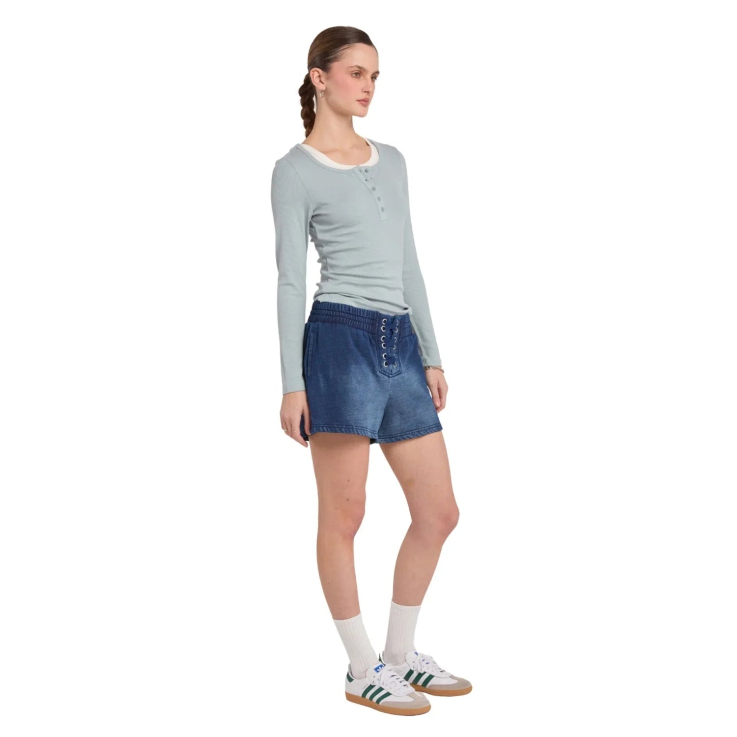 Woman wearing blue denim lace-up shorts, long sleeve top, and sneakers on white background