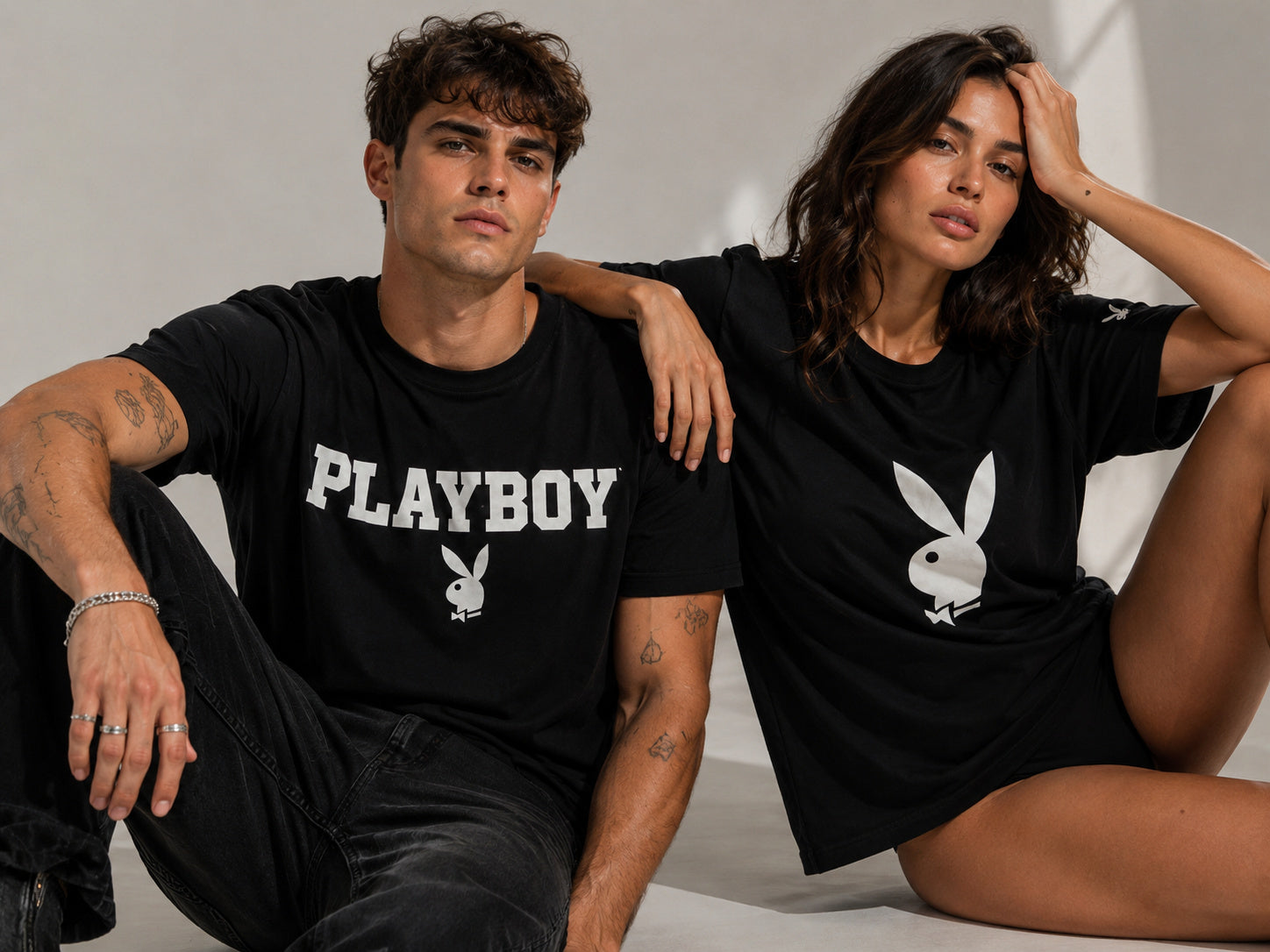 A man and a woman seated side by side against a light seamless backdrop, both wearing black Playboy tees — his with the arch PLAYBOY wordmark, hers with the bunny head.