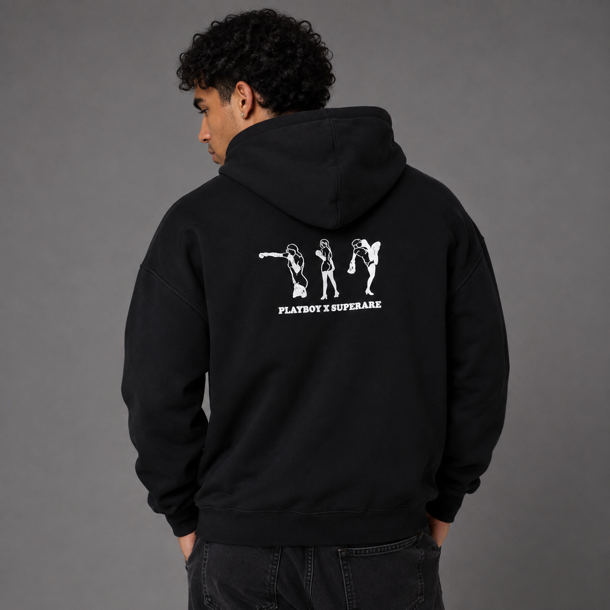 A man wearing a black hoodie from the back, featuring a white graphic print of three dancing figures and the text 'PLAYBOY X SUPERSTAR' on the back, set against a solid gray studio background.