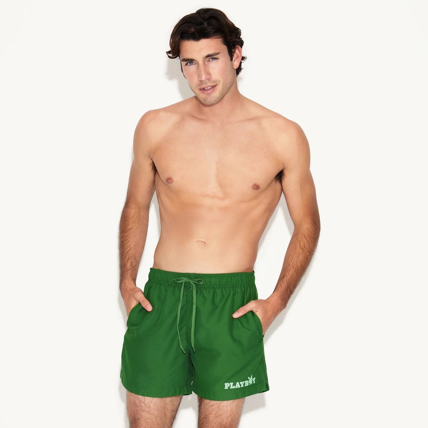 Shirtless man modeling green men's swim trunks with white logo and drawstring waist.