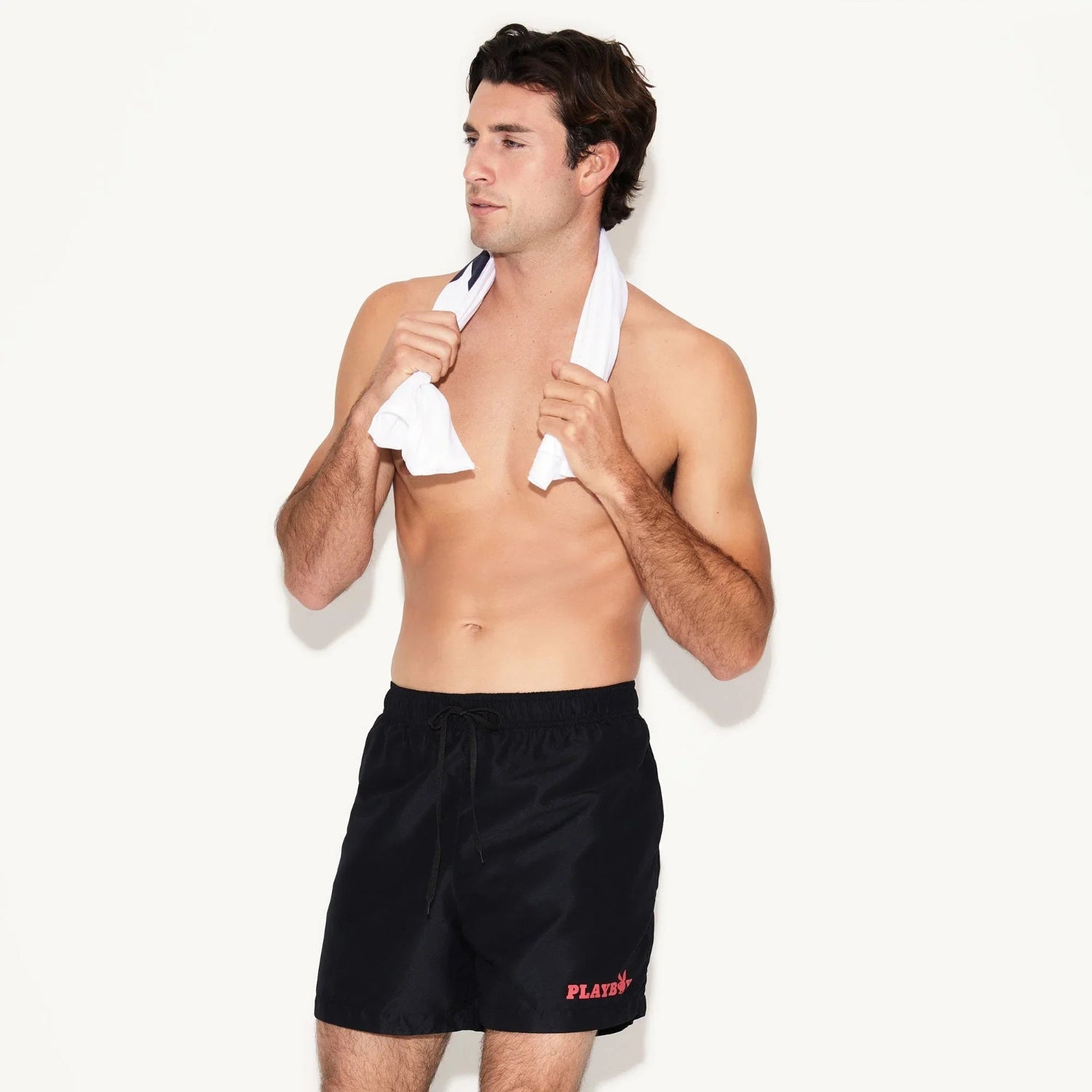 Shirtless man wearing black men's swim trunks with red text detail, holding white towel