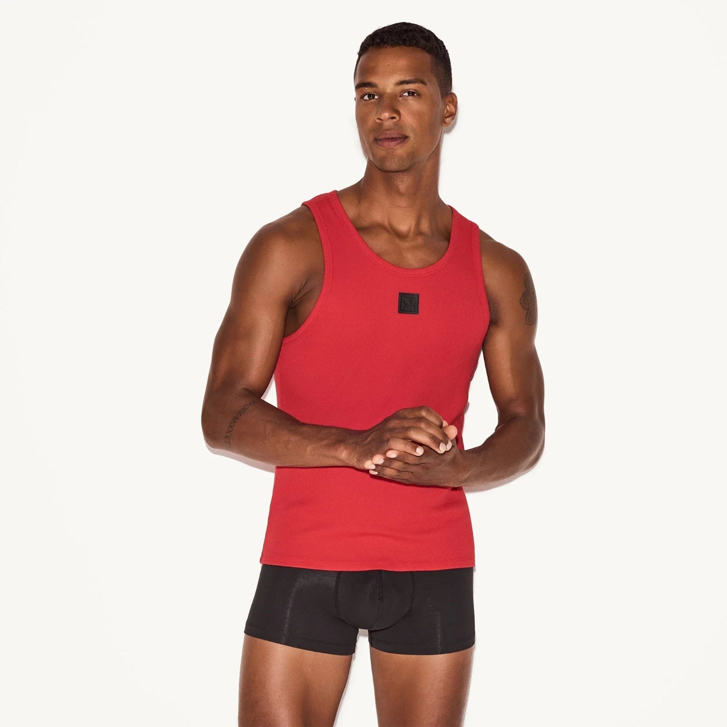 Man wearing a red ribbed tank top and black boxer briefs against a white background