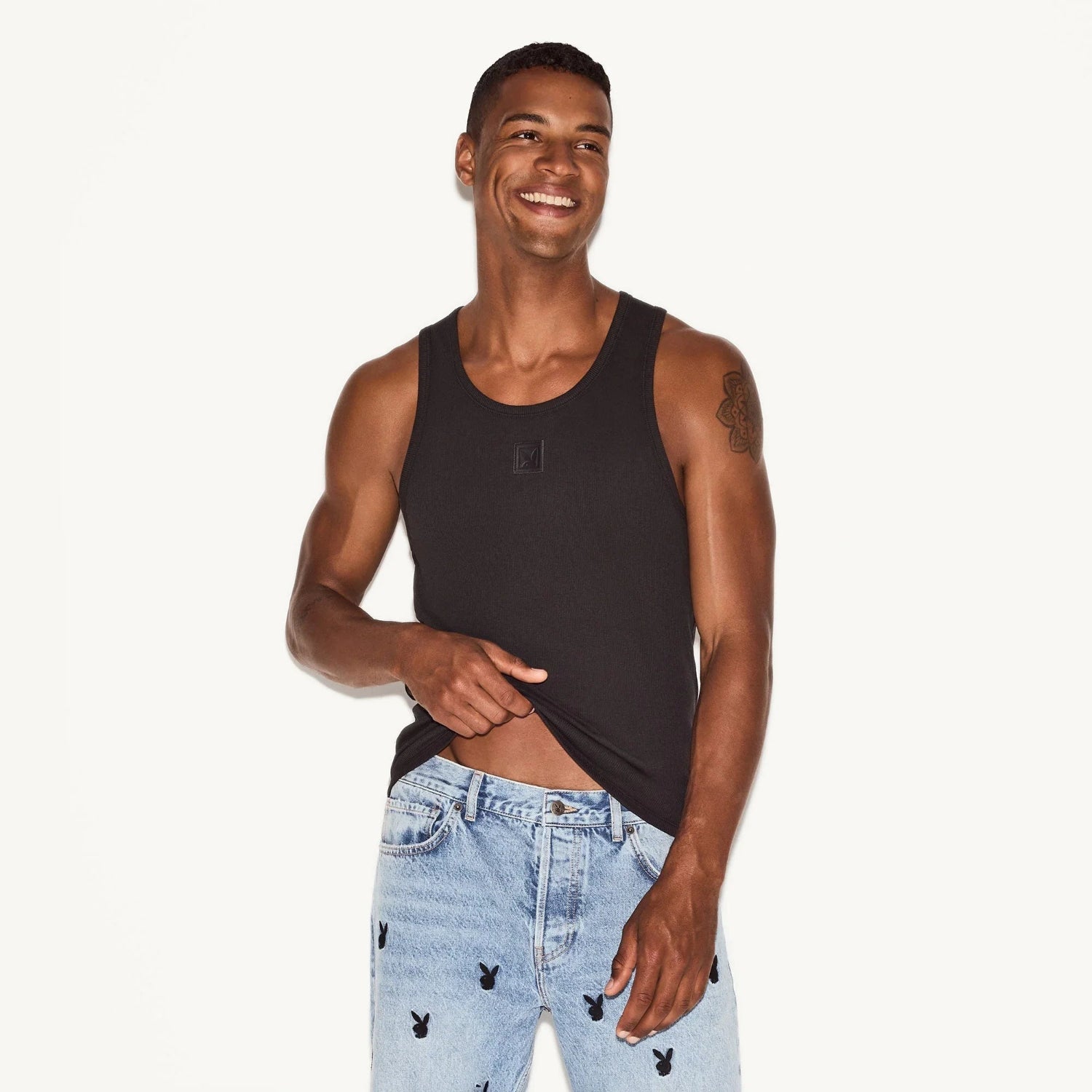 Man in black ribbed tank top and light blue jeans with bunny logo print standing against white background