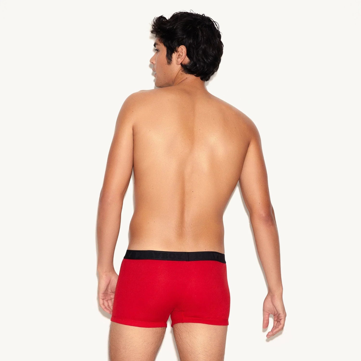 Man wearing red ribbed trunk underwear with black waistband, rear view on light background