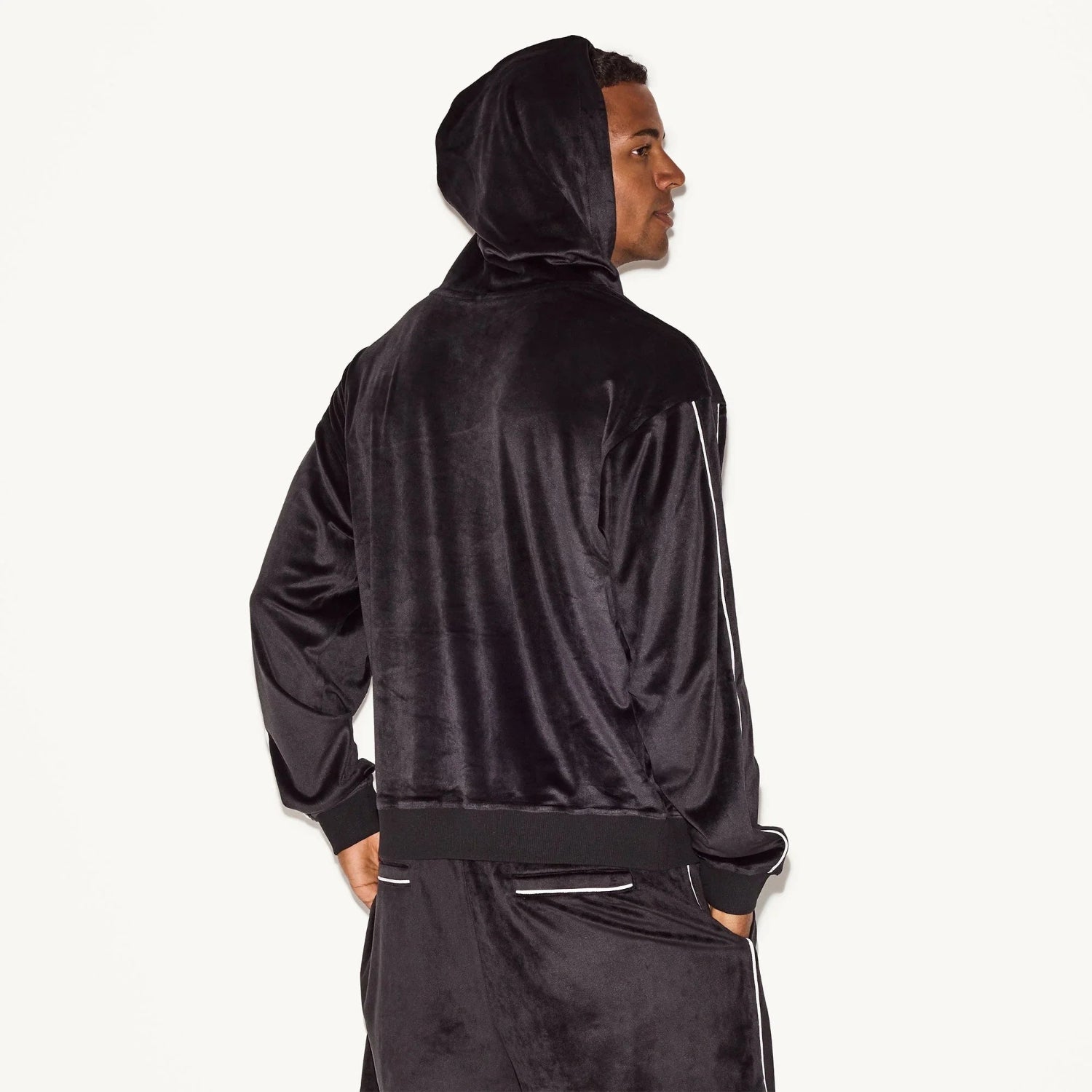 Man wearing black velour hoodie and matching pants, side view, white background