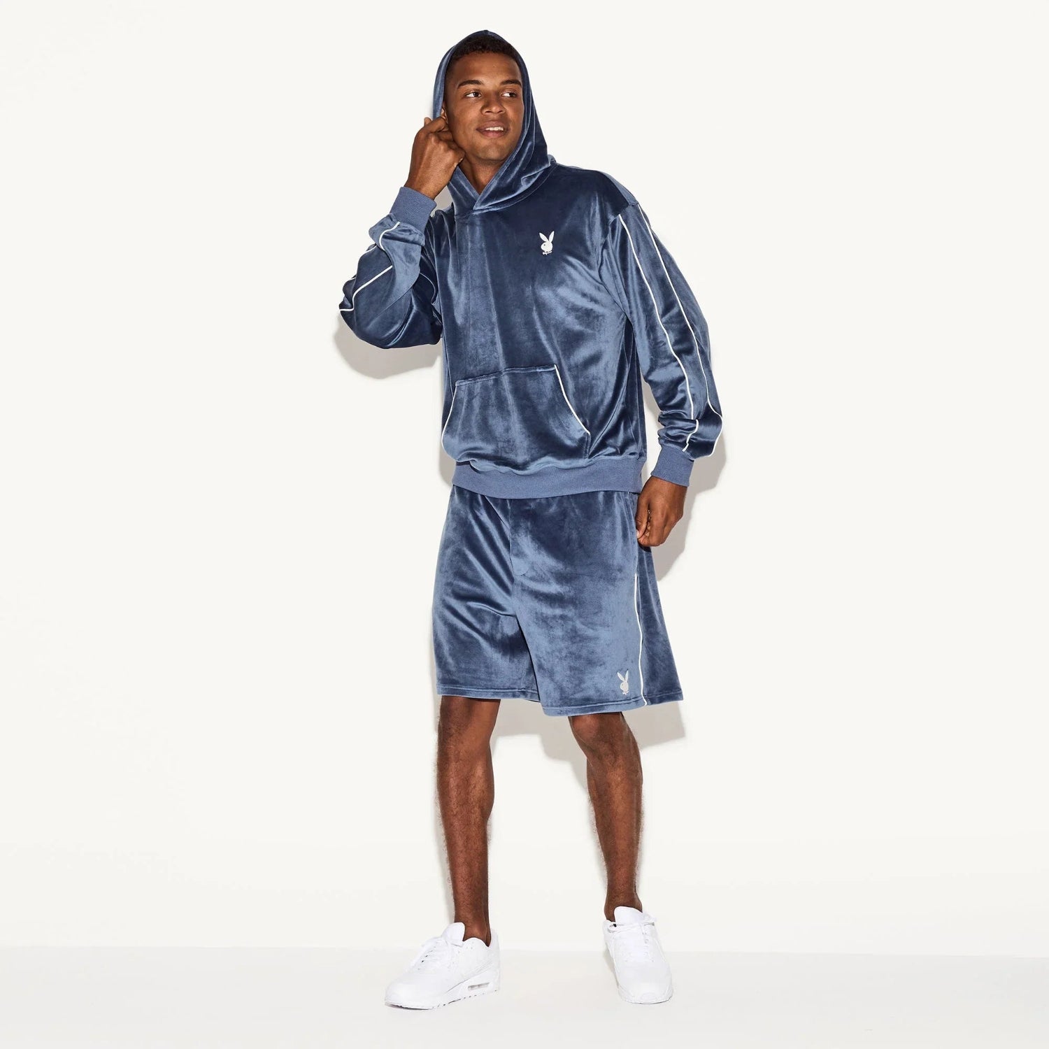 Man wearing blue velour hoodie and shorts set with bunny logo, white sneakers, white background