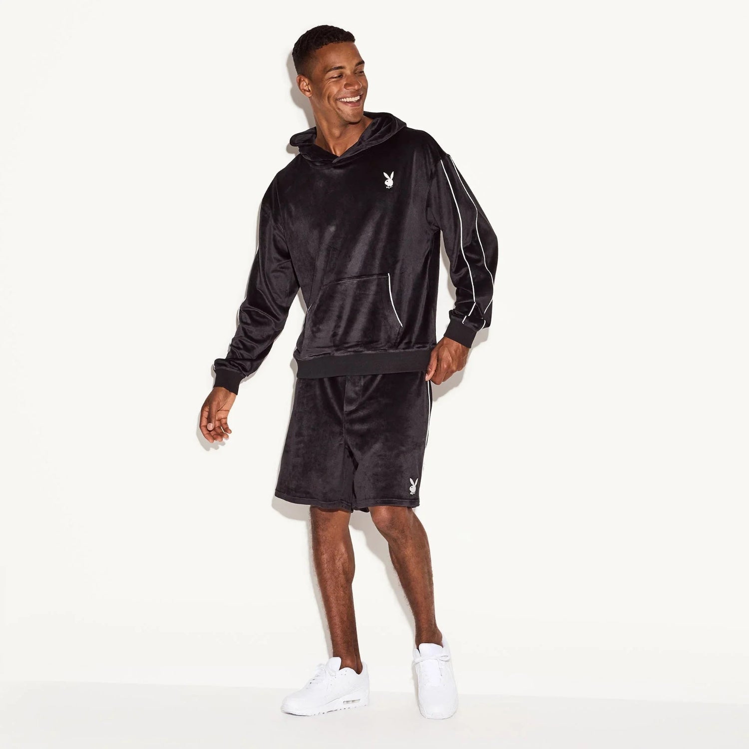 Man wearing black velour hoodie and shorts set with white bunny logo, standing indoors