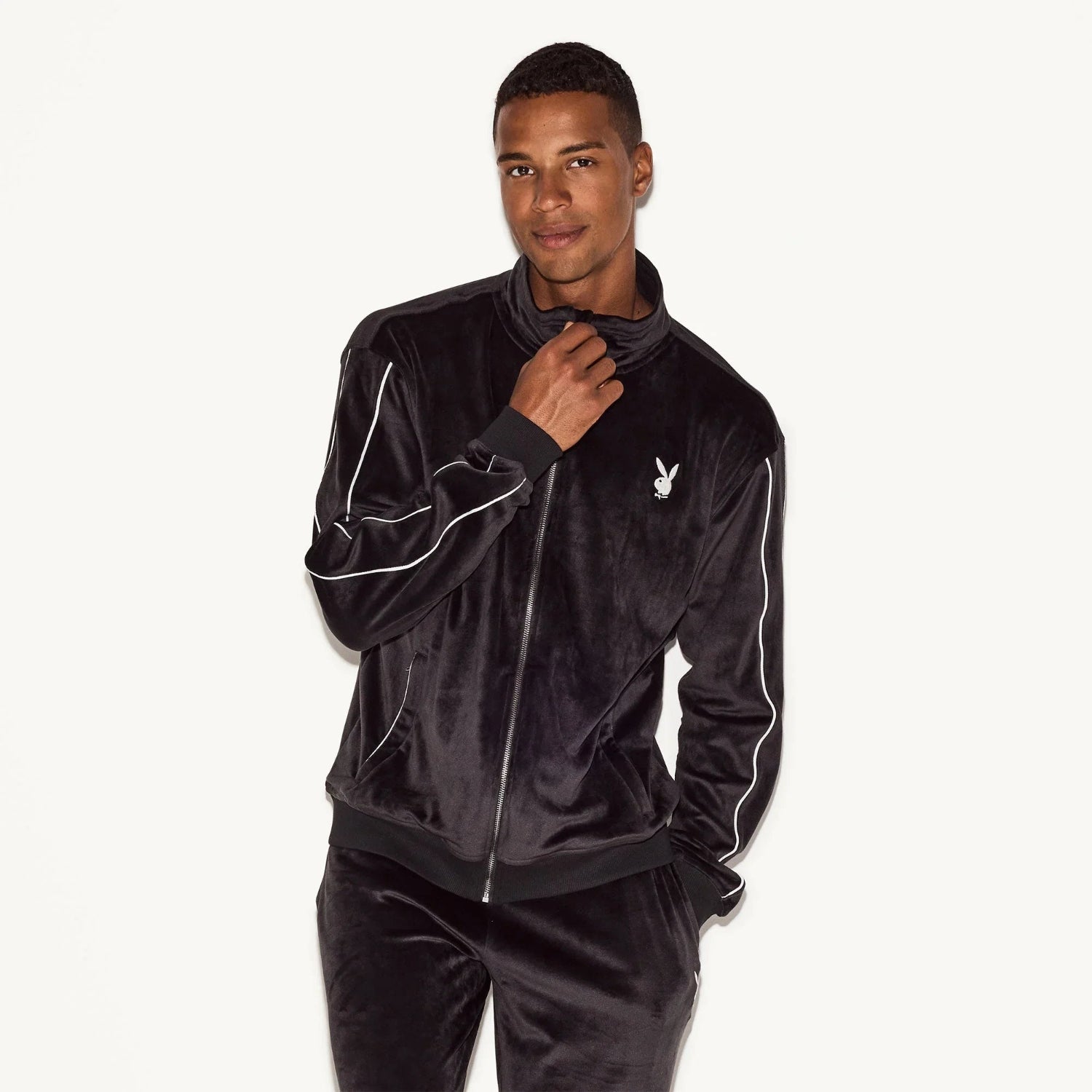 Man wearing black velour zip-up jacket with white piping and bunny logo on chest