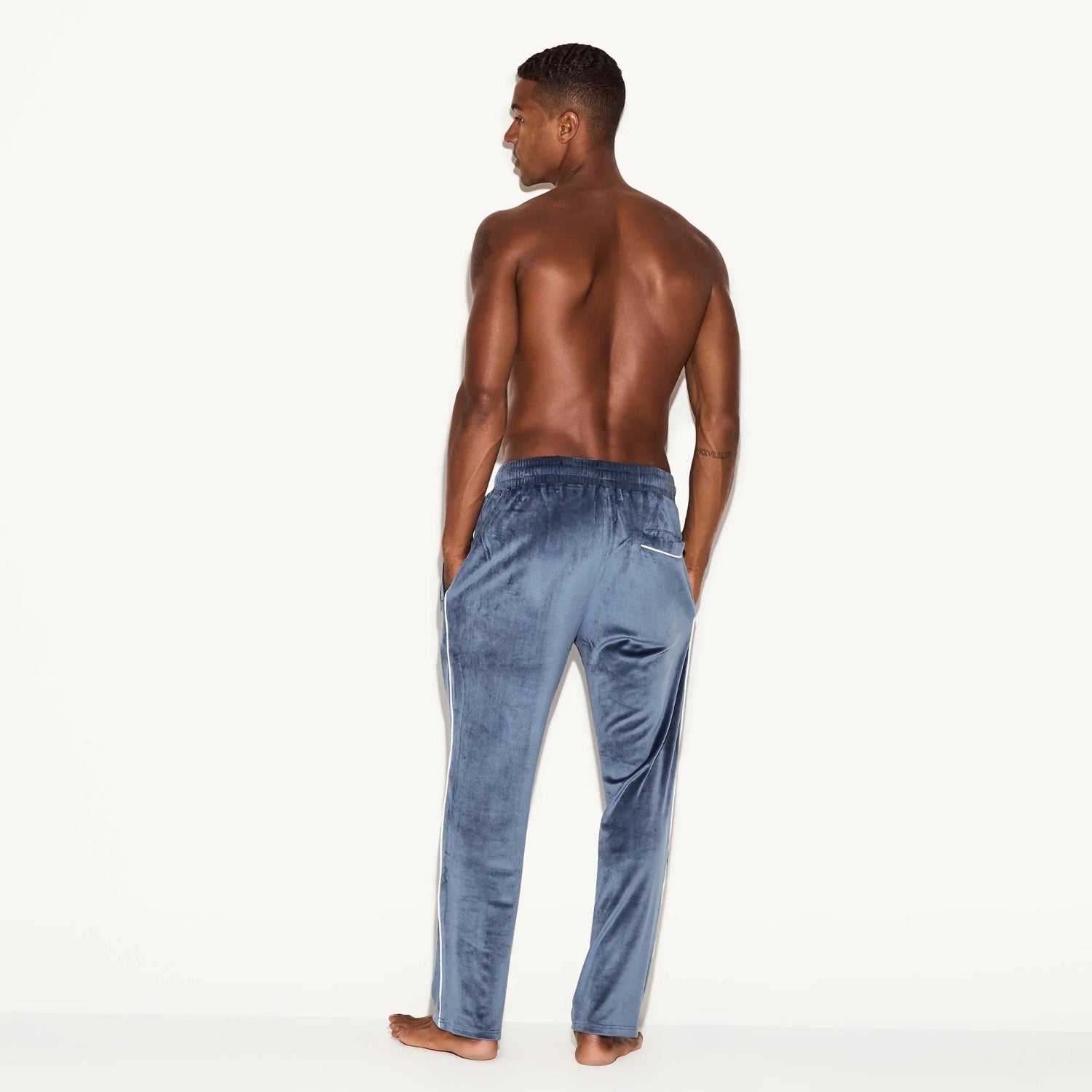 Back view of a man wearing blue velour lounge pants with white trim on a plain background