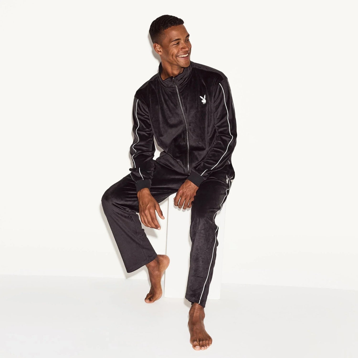 Man wearing black velour track pants and matching zip jacket with white piping and chest logo