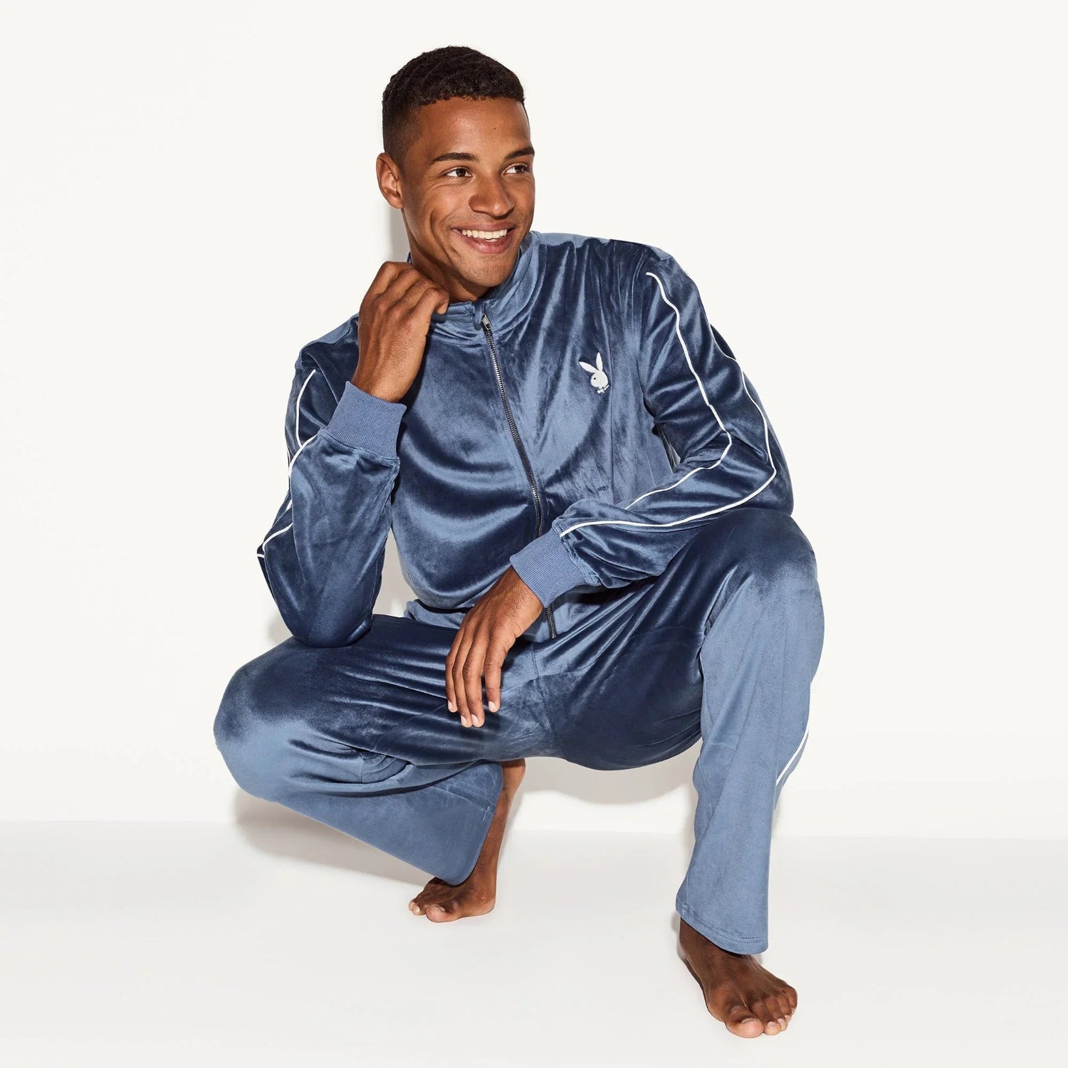 Man wearing blue velour tracksuit with white piping and embroidered bunny logo, squatting barefoot.