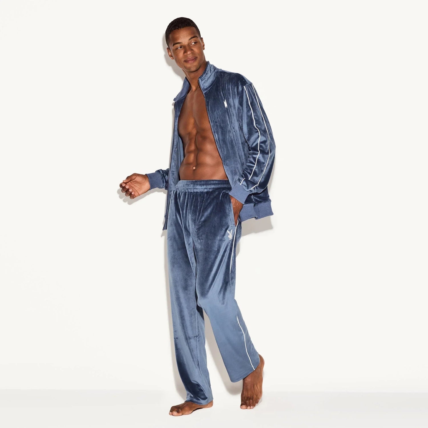 Man modeling blue velour track pants with elastic waistband and white logo on hip