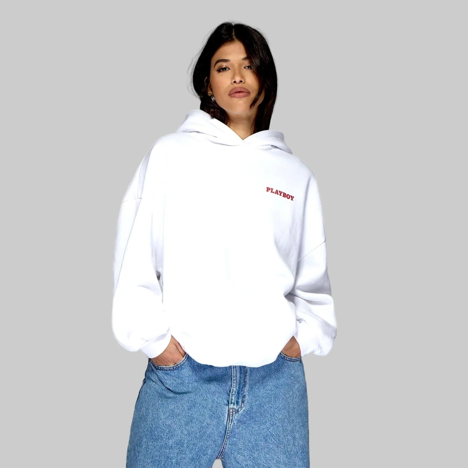 Woman wearing white hoodie with red Playboy logo and blue jeans against gray background