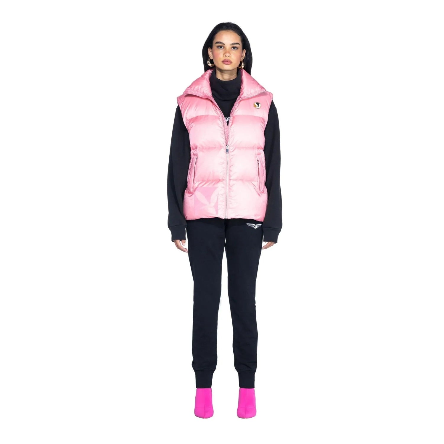 Woman wearing pink puffer vest over black sweatshirt and joggers with pink shoes, studio shot