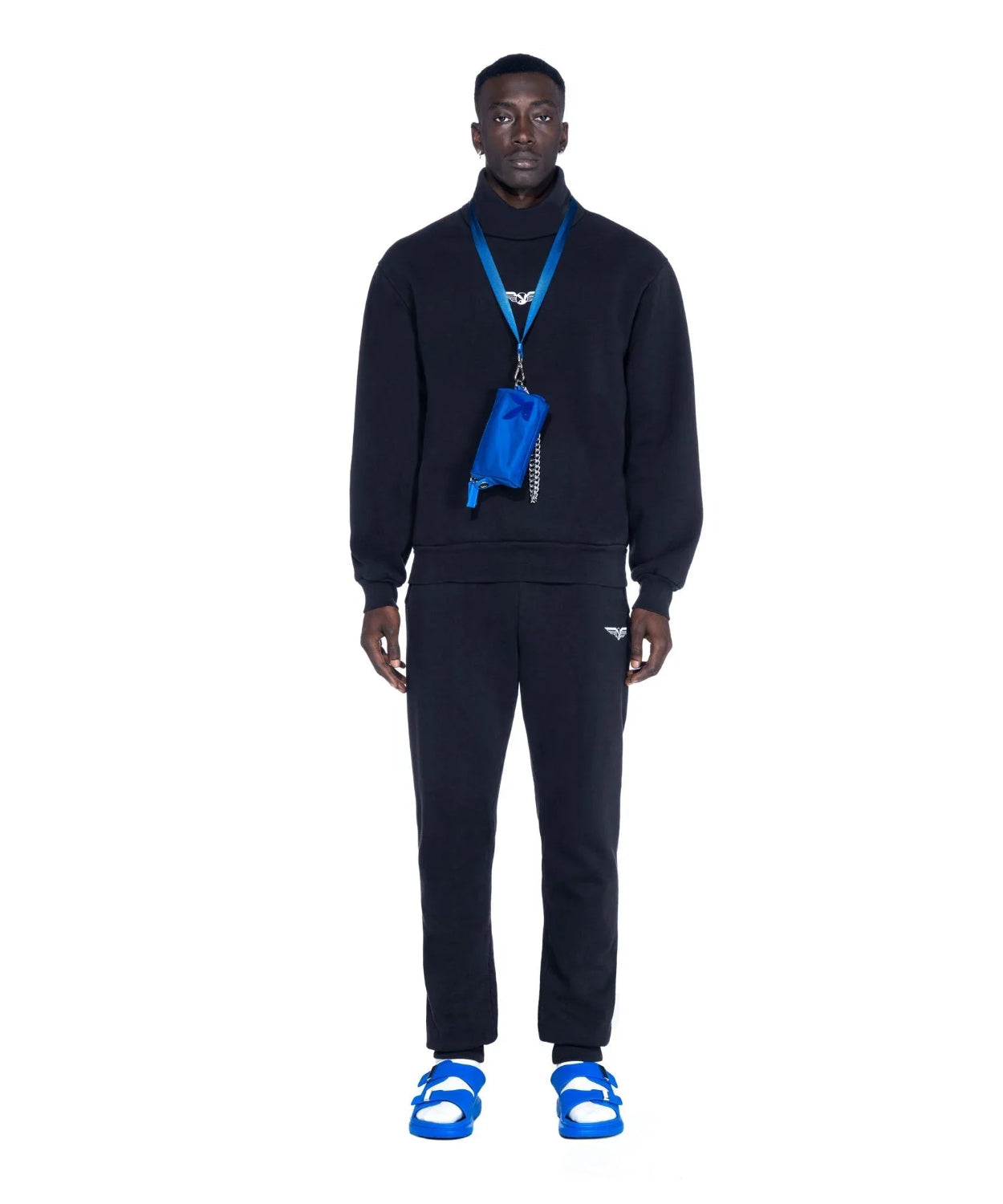 Man wearing black tracksuit, blue sandals, and a blue nylon pouchette crossbody bag.