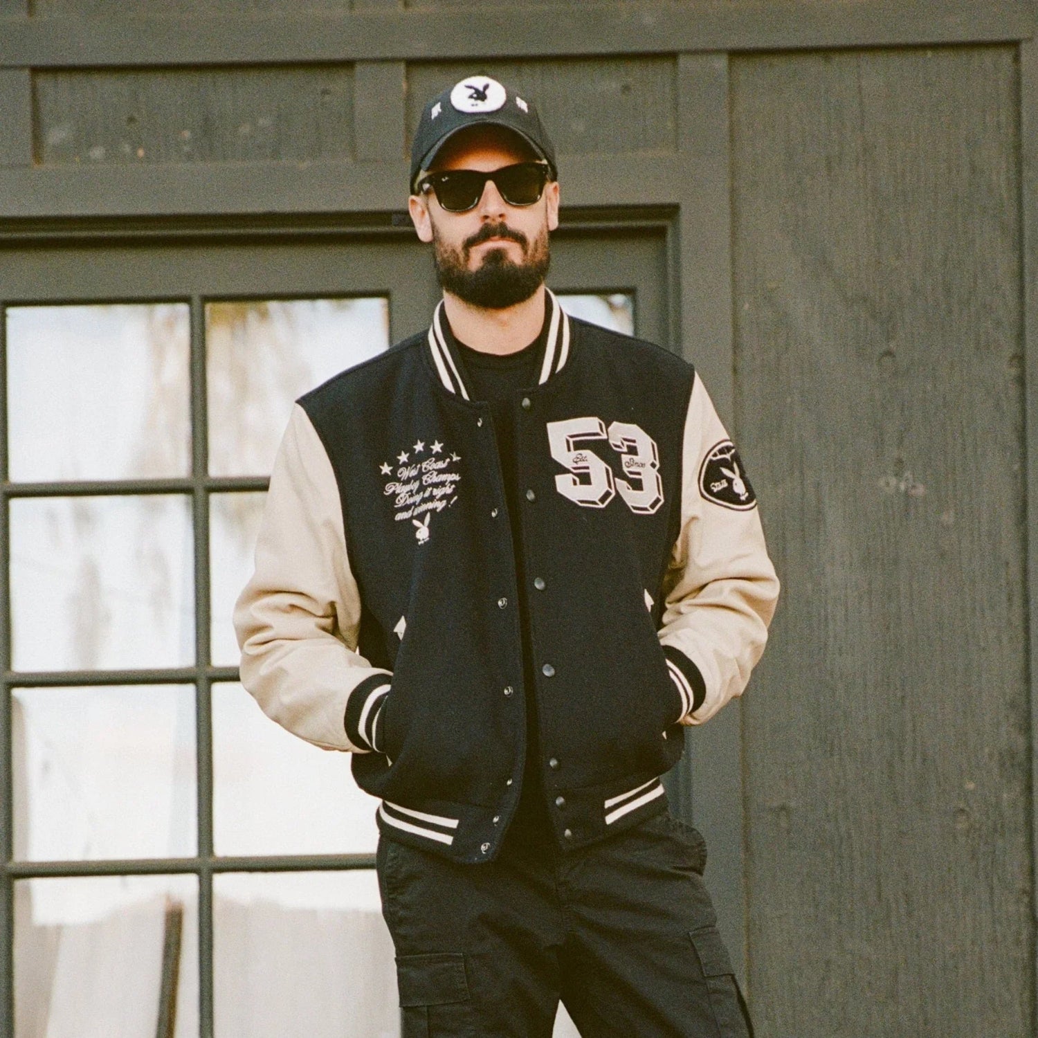 Man wearing black and beige varsity jacket with '53' patch and logo, sunglasses, and cap