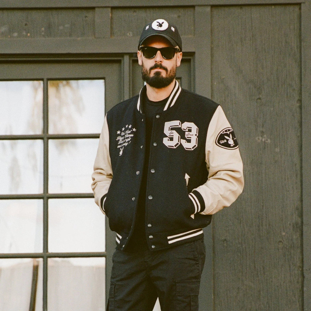 Man wearing black and beige varsity jacket with '53' patch, cap, and sunglasses outdoors