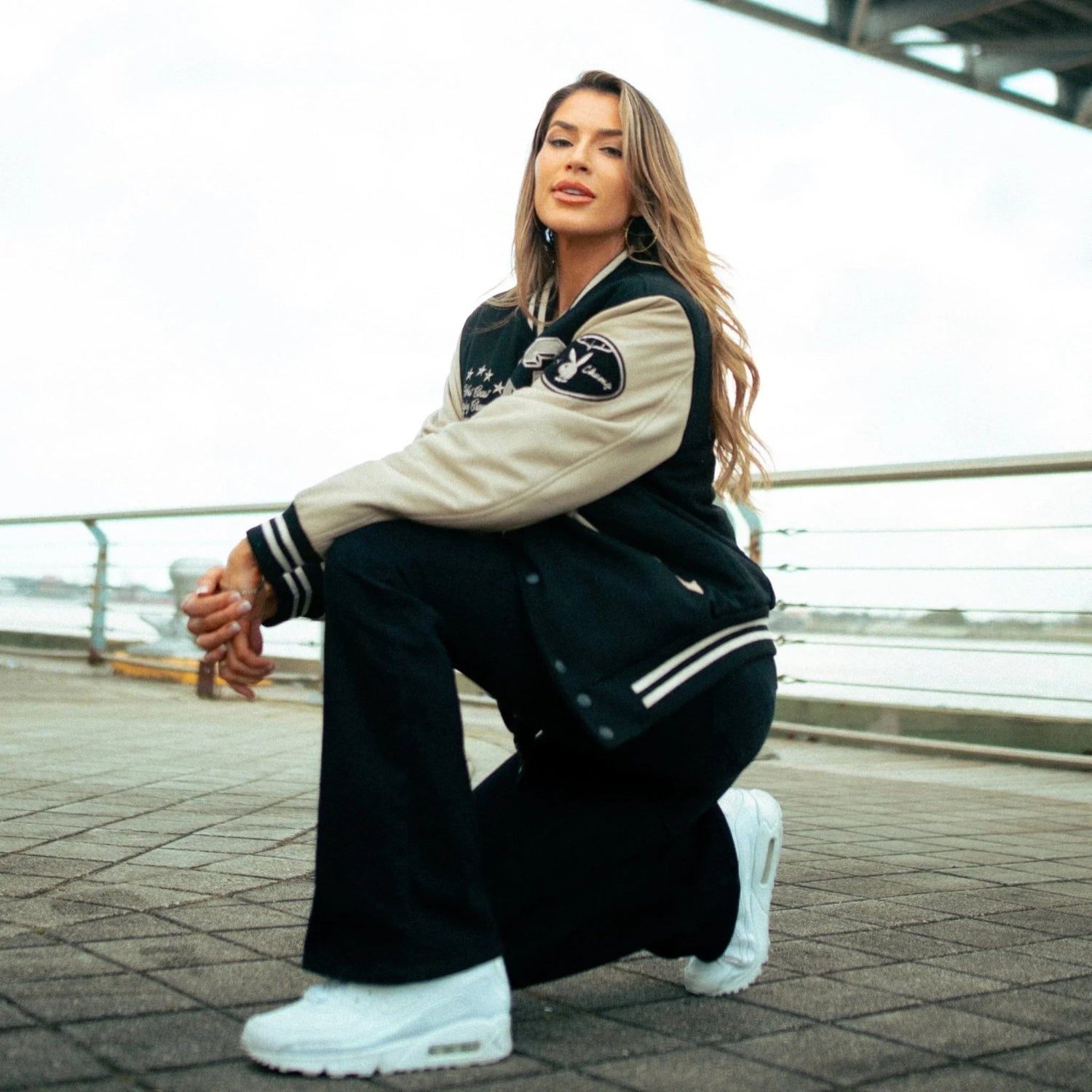 Woman wearing a black and beige varsity jacket, black pants, and white sneakers outdoors.