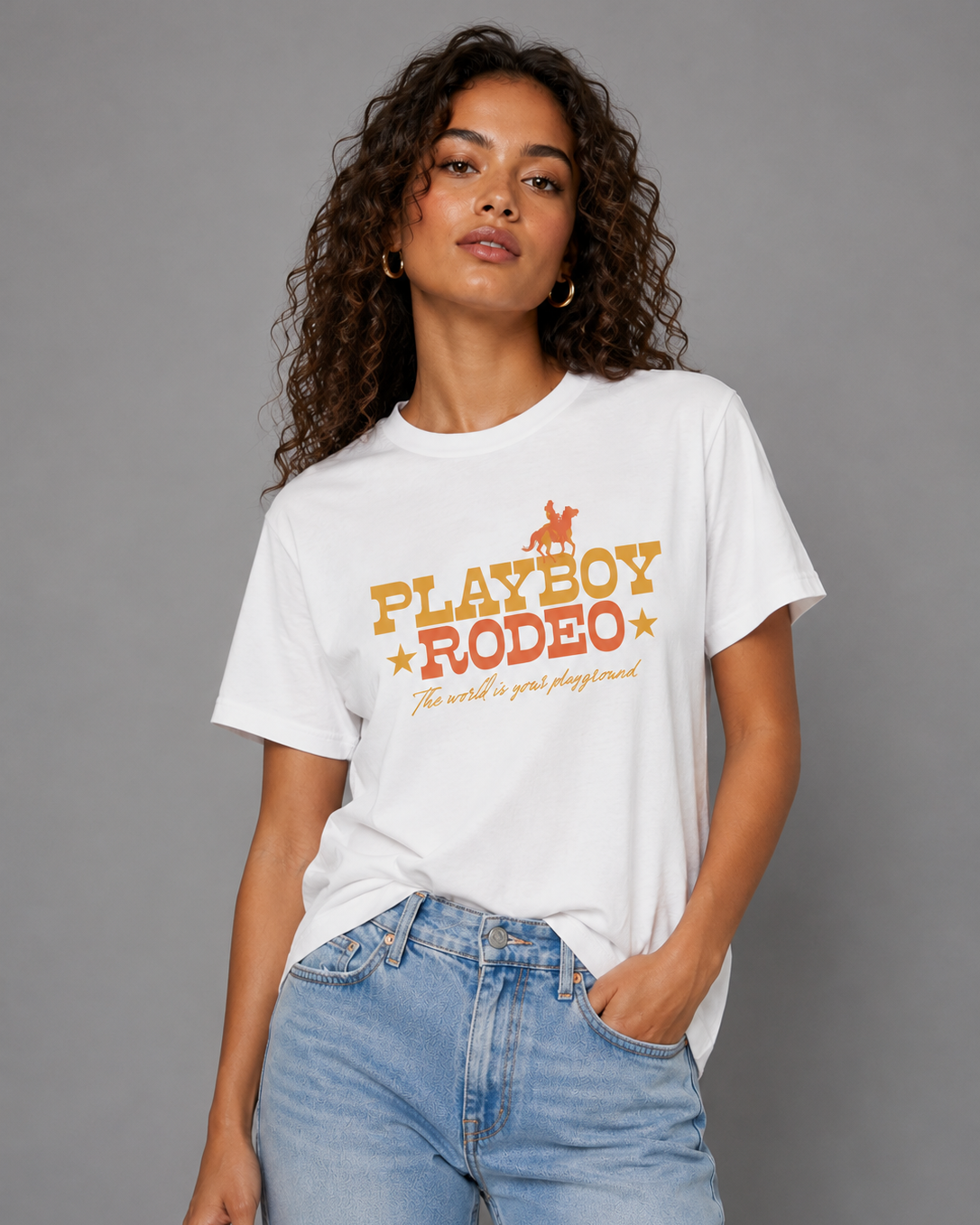 A woman wearing a white short-sleeve graphic T-shirt with 'PLAYBOY RODEO' text and a cowboy silhouette in orange and yellow, paired with light blue jeans, against a plain gray studio background.