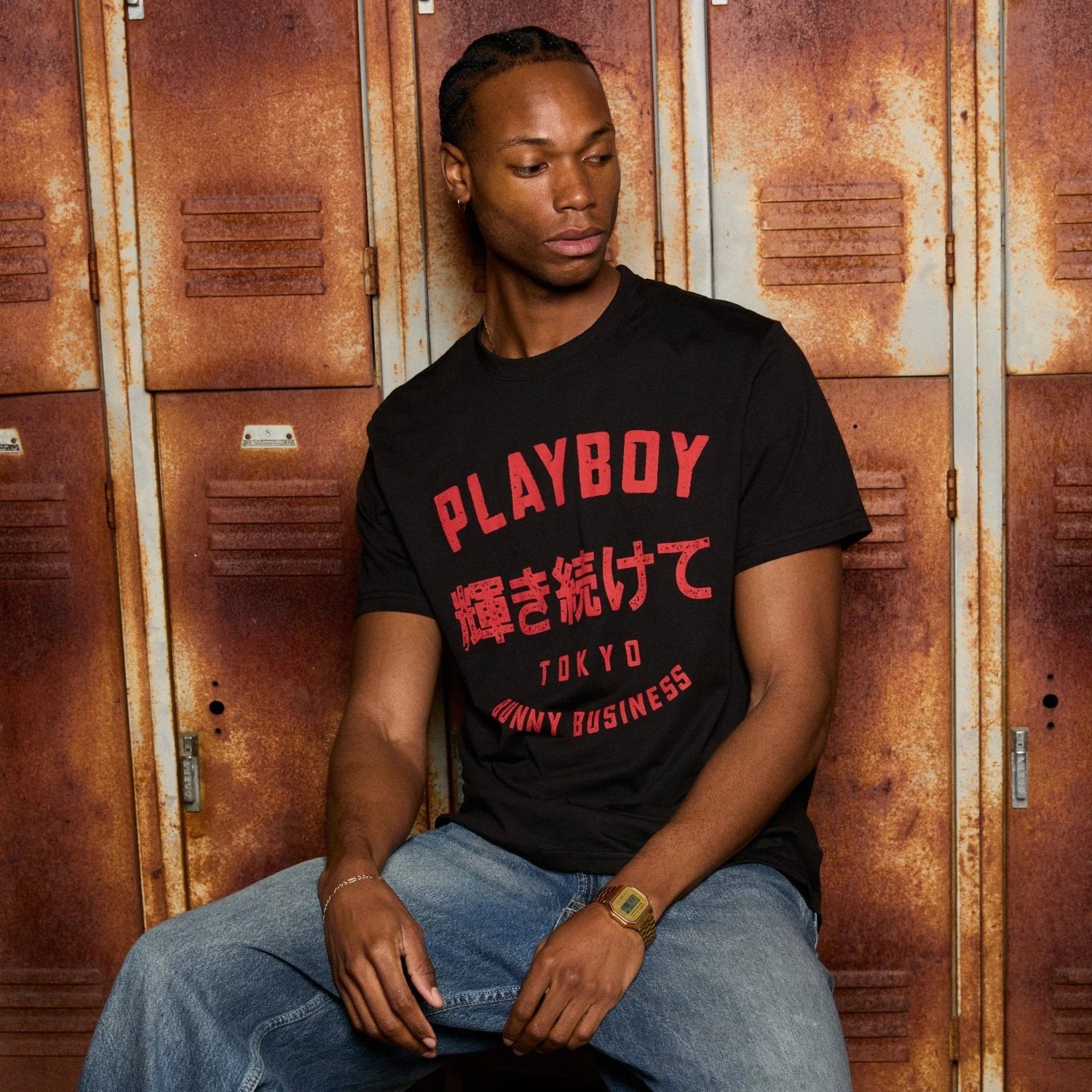 Man in black Tokyo graphic t-shirt and jeans sitting by vintage rusty lockers