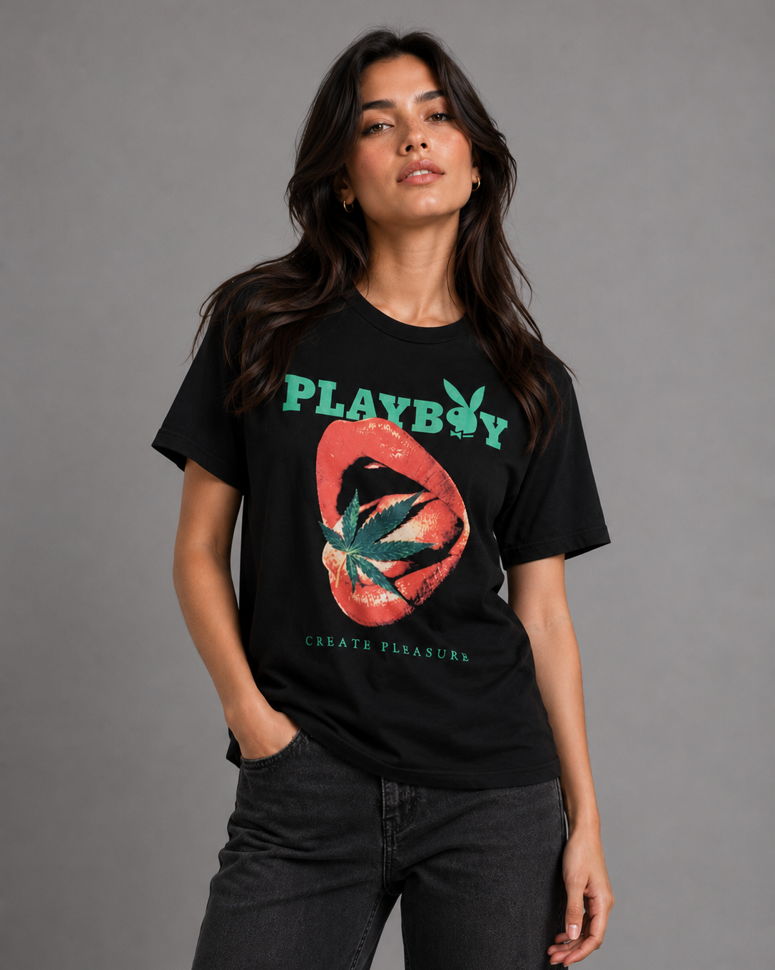 A woman modeling a black short-sleeve graphic t-shirt featuring the green 'PLAYBOY' logo and a red lips-and-marijuana leaf design, paired with dark gray jeans against a solid gray studio background.