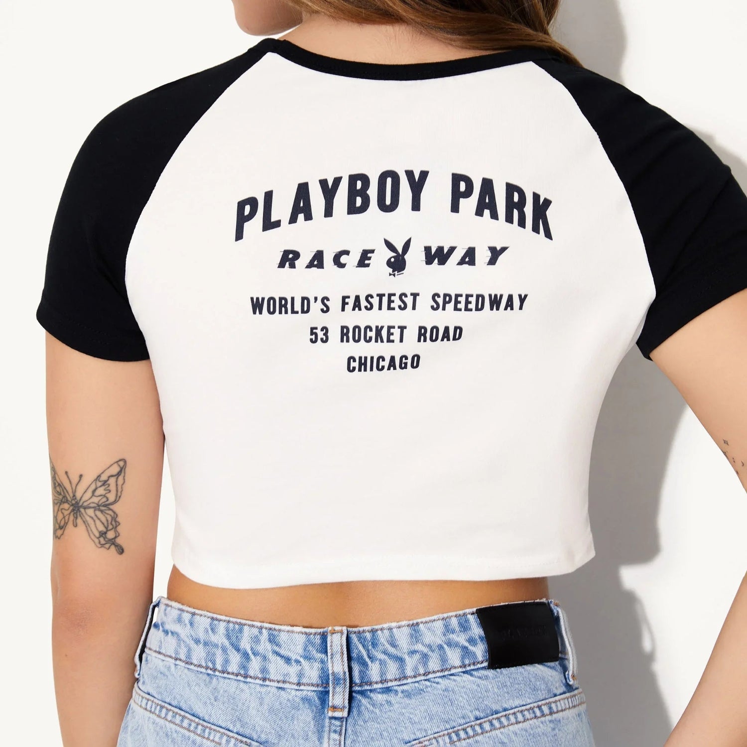 Back view of a woman wearing a white and black crop tee with raceway print and blue jeans